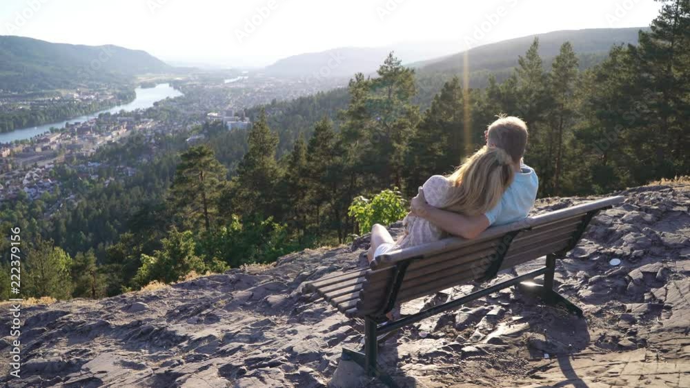 Romantic loving couple sitting on wooden bench and admiring the town and the river in valley, taking selfies and pictures Romantic loving couple sitting on wooden bench and admiring the town and the river in valley, taking selfies and pictures