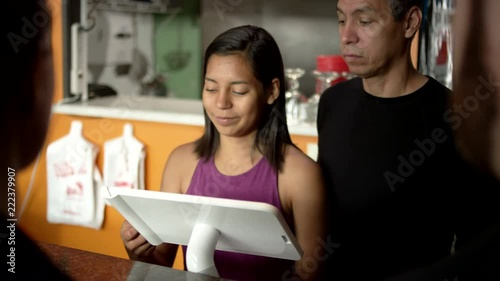 Supportive Hispanic father watches cute teen daughter sweetly take a debit card from customers, swipe it through the touch screen register, and turn the screen around for a signature while practicing the register at their family owned restaurant