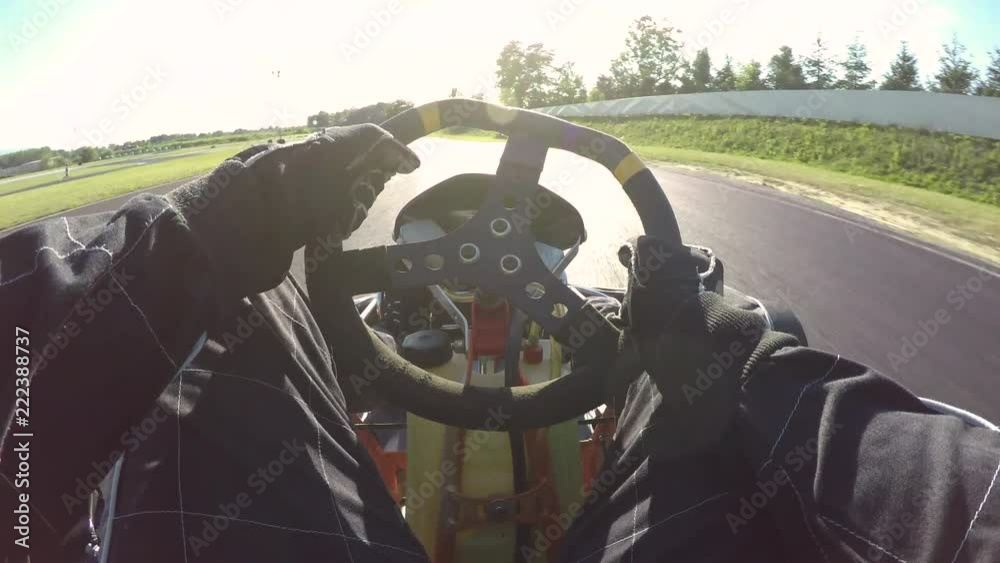 POV, LENS FLARE: Steering a speeding go cart along the winding asphalt circuit on a sunny day. Driving a fast cart in a black suit through sharp turns of a bumpy racetrack. Bright sun shines on racers