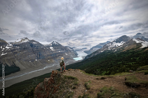 Man hiker on mountain top looking at view of Columbia Icefieds glacier and a moraine lake under stormy sky. Columbia Icefield. Banff / Jasper National Park. Canadian Rocky Mountains. Alberta. Canada.
