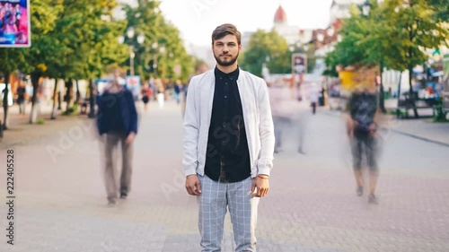 Zoom out time-lapse of attractive guy standing in crowded street in city and looking at camera on summer day while people are moving around. Life and loneliness concept.