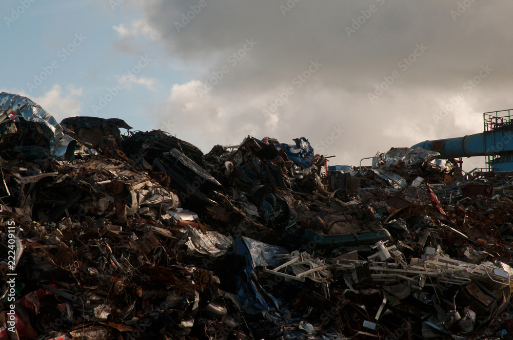 Pile of scrap metal at a junk yard with a metal structure on the side and a cloudy sky in the background