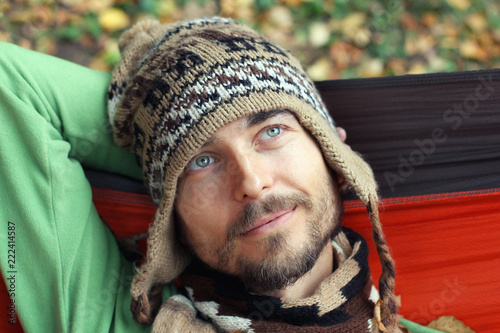 Portrait of a young bearded man in hat and scarf. He is rests in a hammock outdoor. Wistful smile on his face. Autumnl mood concept photo