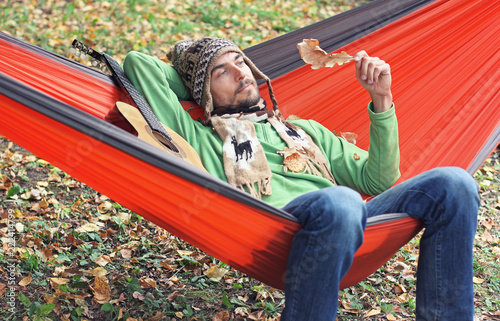 Young bearded man with wistful smile and a guitar relax in a hammock in the autumn forest. Camping life concept