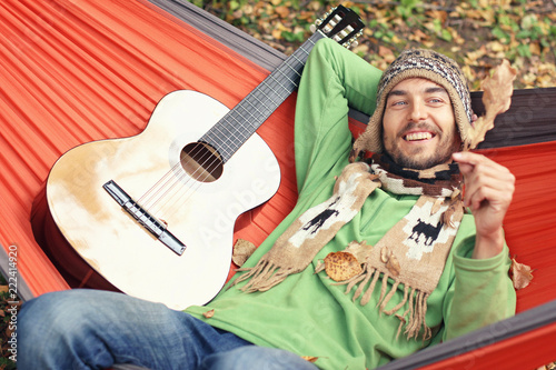 Autumn is coming Young handsome man with guitar relaxs in a hammock outdoor. He smiles and looks on an oak leaf in his hand. Fall mood concept