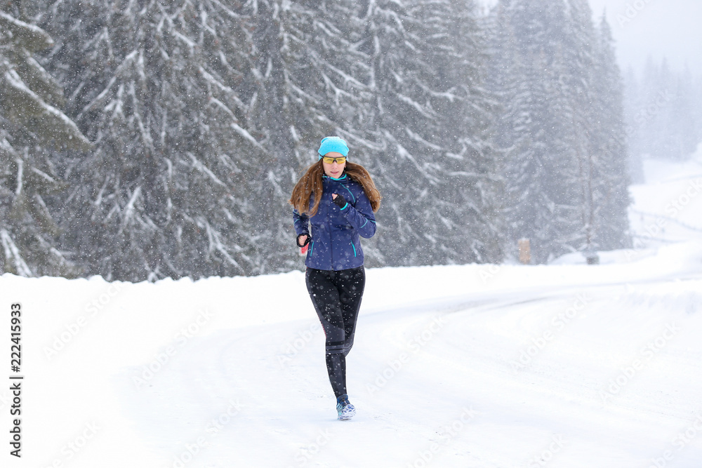 Young woman running on the mountain road in bad weather in winter. Beautiful girl jogging in blizzard or snowstorm