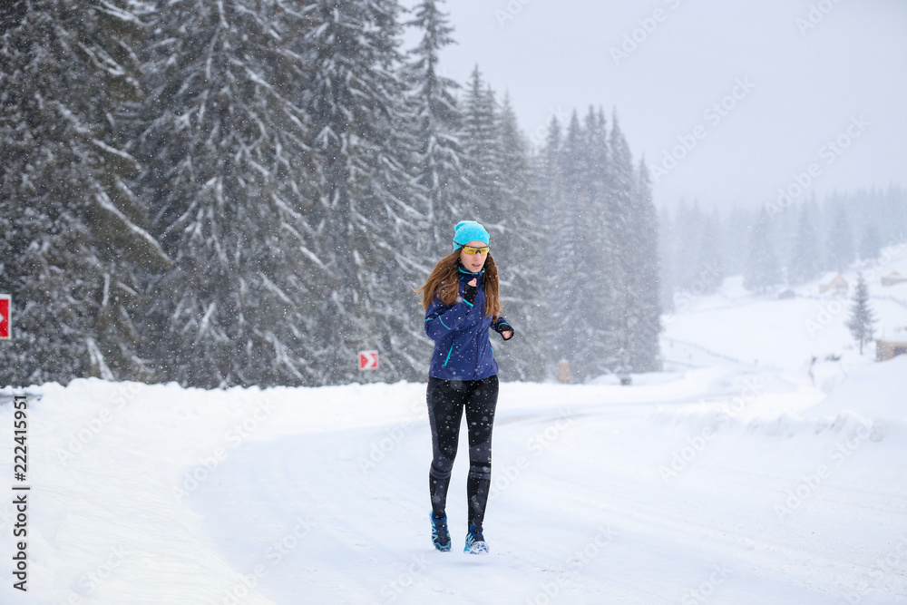 Young woman running on the mountain road in bad weather in winter. Beautiful girl jogging in blizzard or snowstorm