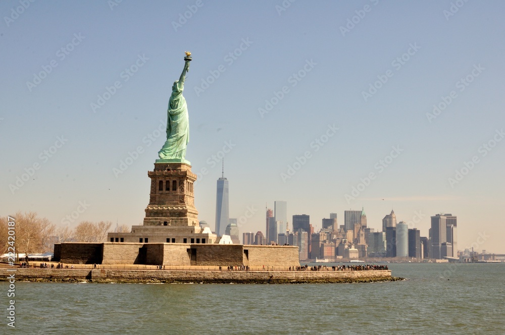 Statue de la liberté à Ellis Island au premier plan vue de la mer avec l'île de Manhattan  au fond par une matinée ensoleillée et un ciel bleu à New York, Etats-Unis