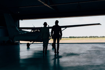 rear view of silhouettes of stylish couple walking near airplane in hangar