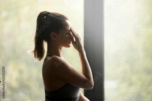 Young sporty woman practicing yoga, doing Alternate Nostril Breathing exercise, nadi shodhana pranayama pose, working out, wearing sportswear, grey top, indoor close up, yoga studio, side view