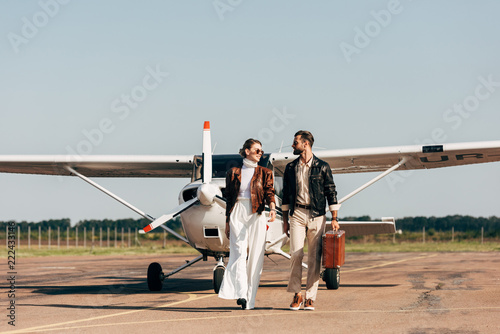 stylish young couple in leather jackets and sunglasses walking with retro suitcase near airplane photo