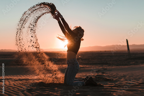 Side view of young woman kneeling touching and throwing the sand in a desert sunset background