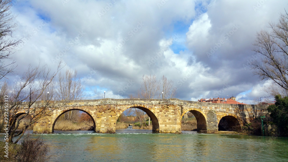Fototapeta premium View on the stone ancient bridge over the river Cea in the vicinity of the city Sahagun, Spain. Silent river, sunny day, cloudy sky.