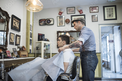 Barbero moderno con vestimenta vintage corta el pelo a joven muchacho veiteañero en barbería.