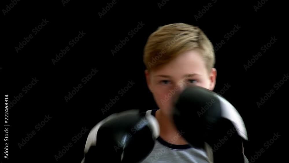 A teenage athlete stands on a dark background and holds his hands in gloves. The boy almost covers his face, his eyes are visible. He has a gray shirt without sleeves, he has blonde hair. A teenage athlete stands on a dark background and holds his hands in gloves. The boy almost covers his face, his eyes are visible. He has a gray shirt without sleeves, he has blonde hair.