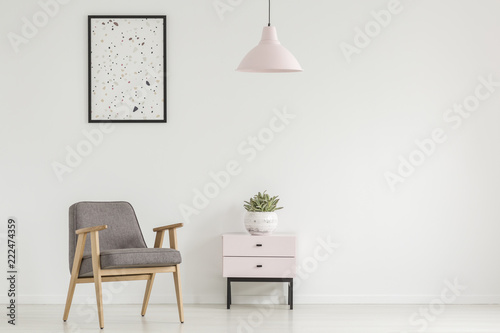 Poster above grey wooden armchair next to cabinet with plant in white flat interior with lamp. Real photo © Photographee.eu