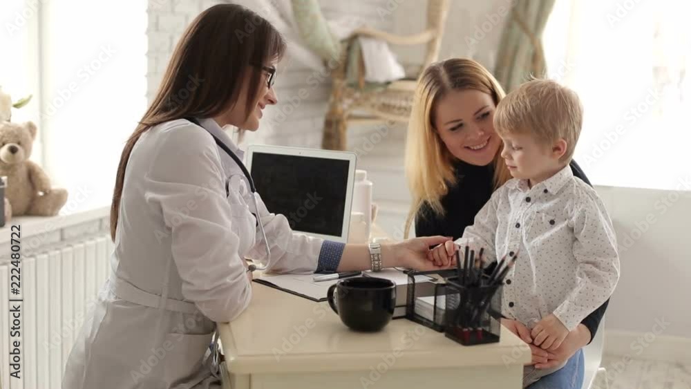 Kind and sweet woman doctor talking with a little boy in a children's clinic. Mom with a little son at a pediatrician, the doctor is very nice talking with the baby. Little boy is afraid of the doctor Kind and sweet woman doctor talking with a little boy in a children's clinic. Mom with a little son at a pediatrician, the doctor is very nice talking with the baby. Little boy is afraid of the doctor