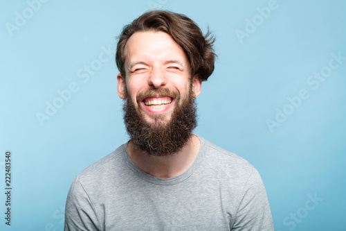 happiness enjoyment and laugh. exhilarated man with a wide grin. portrait of a young bearded hipster guy on blue background. emotion facial expression. feelings and people reaction. photo