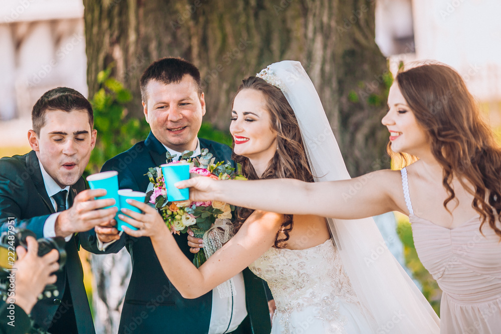 Newlyweds and their friends open champagne bottle standing in the park.  Before wedding ceremony friends having fun and drink champagne. Funny  wedding moment of newlyweds and bridesmaids and groomsmen. Stock Photo |
