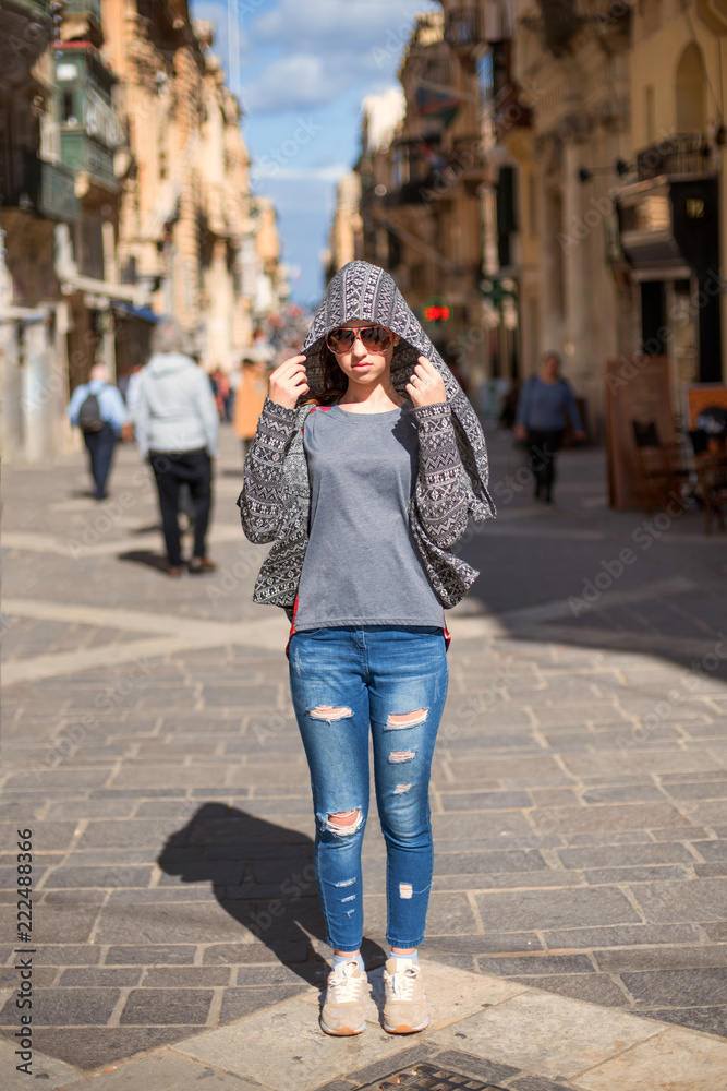 girl tourist in jeans with glasses and a hood in the form of a cobra stands alone in the middle of narrow streets of an ancient city
