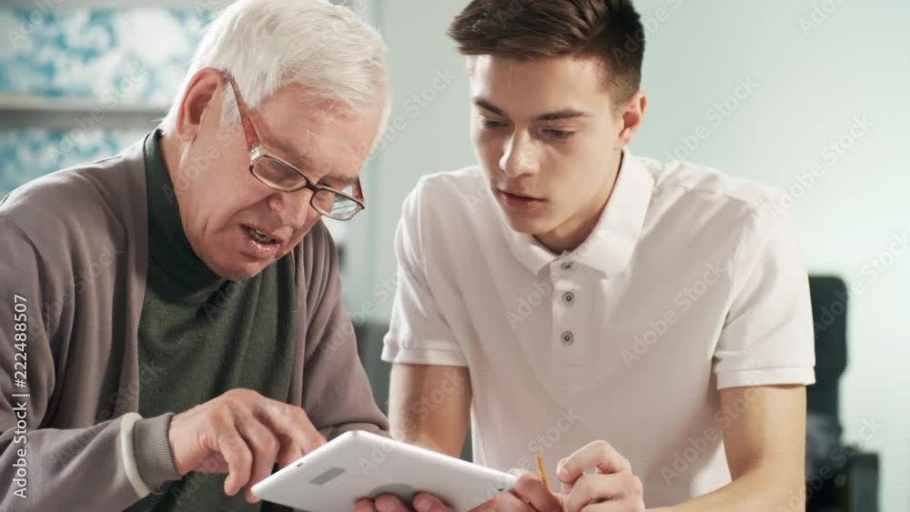 Medium shot of senior man in glasses learning how to use tablet computer application, young caregiver helping him Medium shot of senior man in glasses learning how to use tablet computer application, young caregiver helping him