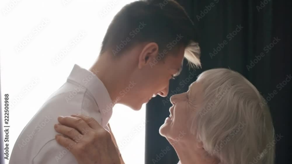Tilt down medium shot of caring grandmother talking to grandson or son, hugging and kissing him, side view