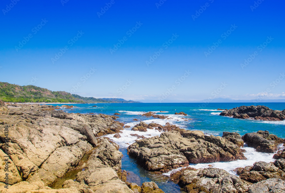 Beautiful view of beach with rock formations in the ocean with waves approaching and blue sky near sand and forest