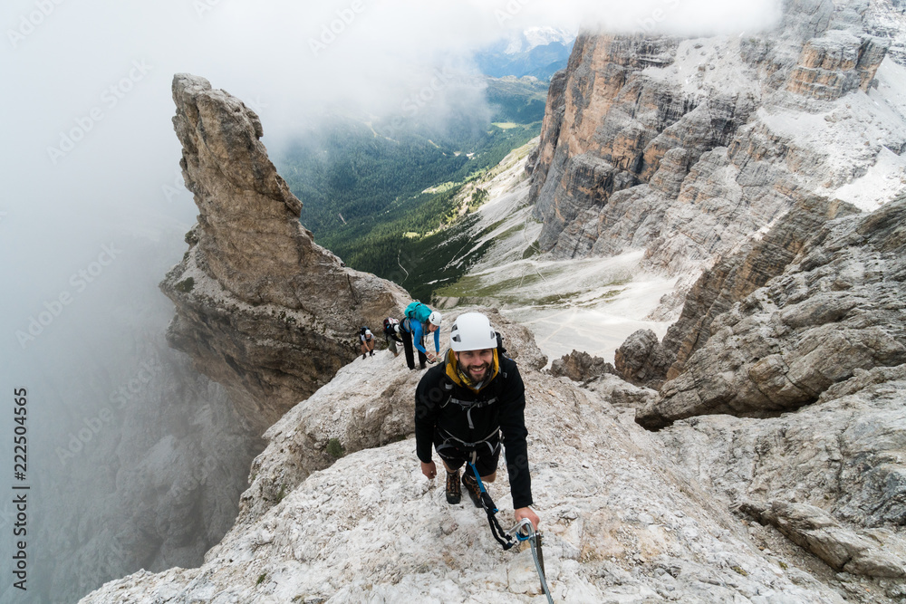 group of young mountain climbers on a steep Via Ferrata with a grandiose view of the Italian Dolomites in Alta Badia behind them