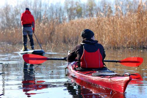 Man paddles a red kayak and man standing with a paddle on the stand up paddle board (paddleboard, SUP) on the river or lake in fall season. Autumn and winter kayaking