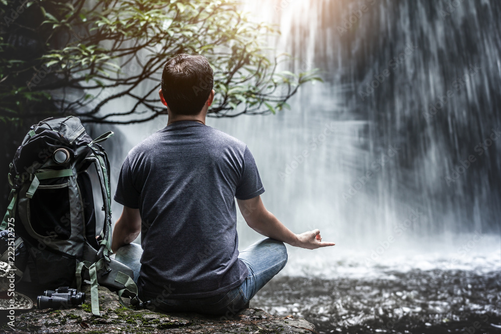 Obraz premium Yoga of traveler relaxing in front of waterfall with backpack and travel equipment. Travel lifestyle concept
