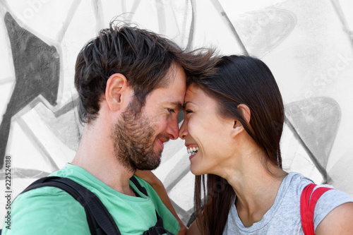 Interracial couple young happy Asian woman and Caucasian man lovers smiling laughing kissing portrait. Tourists on travel holiday taking phone selfie picture hugging each other.