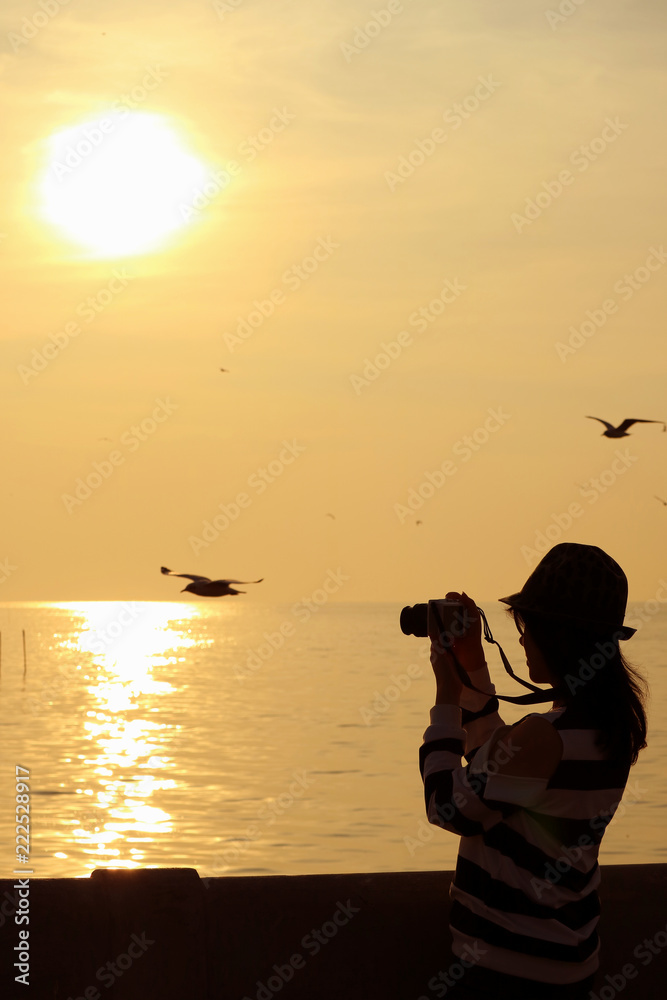 Silhouette of a woman taking pictures of flying seagulls during the sun rising on golden sky over the Gulf of Thailand 