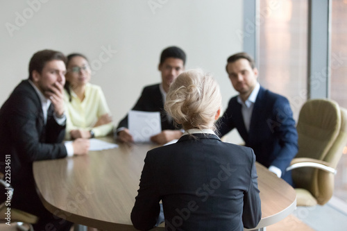 Woman passing job interview at office. Candidate sitting her back to camera. On a background black boss, director holding resume paper and young multiracial colleagues. Interview, hiring, hr concept