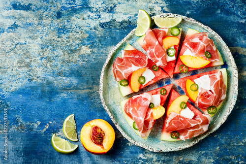 Fresh summer watermelon pizza with feta cheese, peach, prosciutto, jalapeno and honey drizzle on blue background
