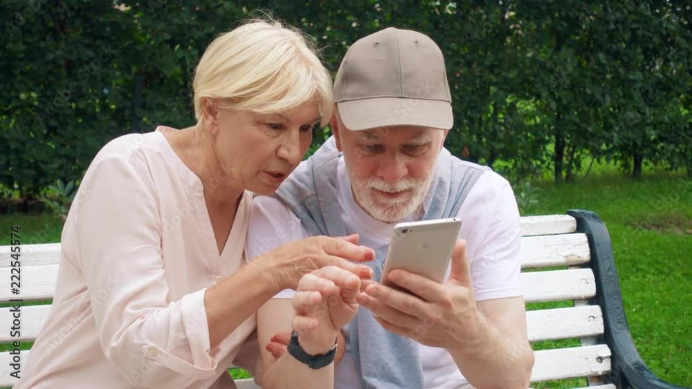 Senior couple having great time sitting on bench in park chatting relaxing, browsing in smart phone. Happy family of retired pensioners enjoying summer vacation outdoors Senior couple having great time sitting on bench in park chatting relaxing, browsing in smart phone. Happy family of retired pensioners enjoying summer vacation outdoors