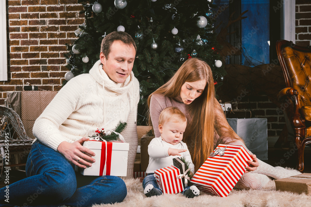 Family celebratory Christmas evening. Family three Caucasian people sitting under coniferous tree Christmas tree on floor of carpet wool. A child holds box with gift, parents young stylishly dressed