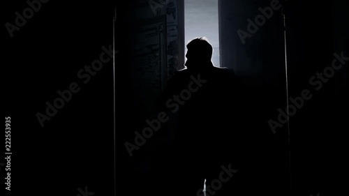 Silhouette of mature man in long dark coat with straight collar opening two massive antique wooden doors revealing empty room in daylight with white dusty walls and big arched windows.