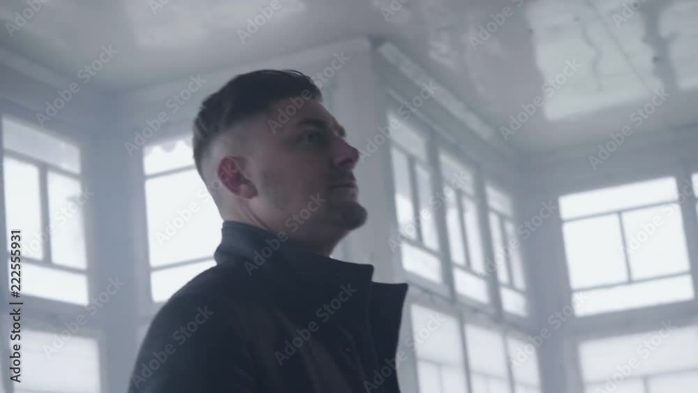 Handsome bearded young man with grey eyes and stylish haircut dressed in classic darken coat with straight collar is looking around himself in empty cold light room surrounded by several windows. Handsome bearded young man with grey eyes and stylish haircut dressed in classic darken coat with straight collar is looking around himself in empty cold light room surrounded by several windows.