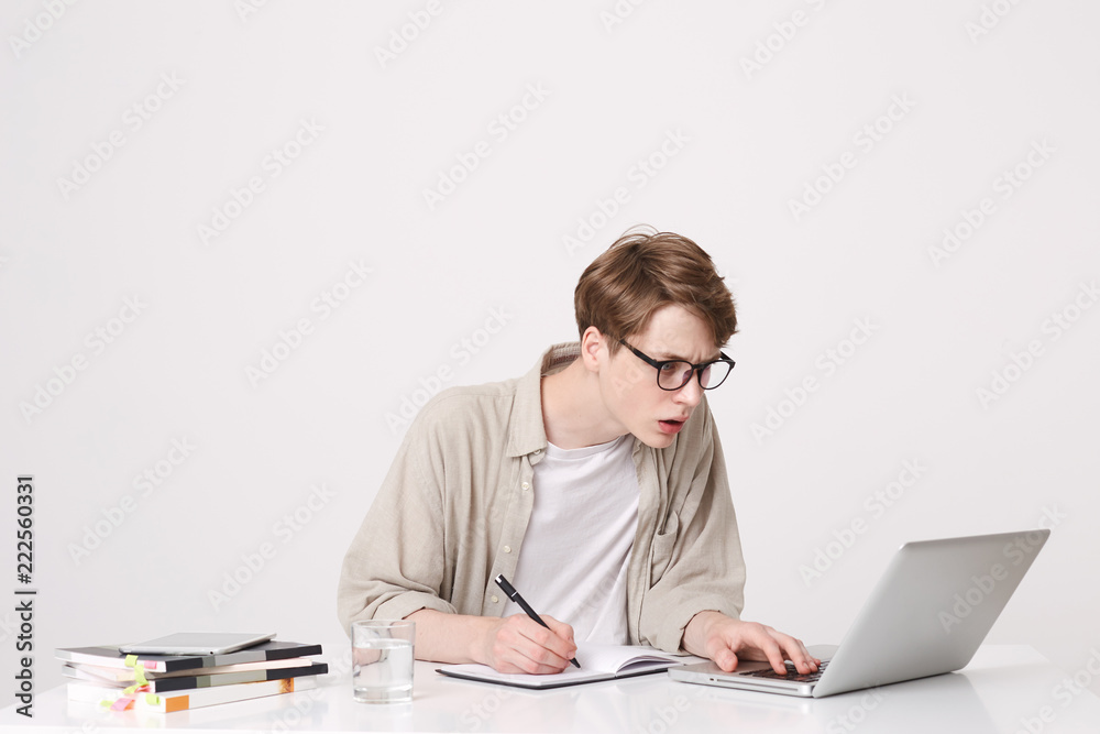 Closeup of concentrated young man student wears glasses and beige shirt sitting and working with laptop computer and notebooks at the table isolated over white background Closeup of concentrated young man student wears glasses and beige shirt sitting and working with laptop computer and notebooks at the table isolated over white background