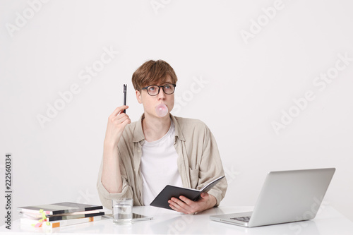 Portrait of pensive young man student wears beige shirt and glasses thinking and blowing bubbles with chewing gum at the table with laptop computer and notebooks isolated over white background
