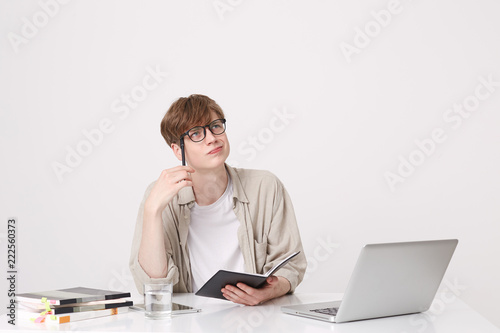 Closeup of pensive handsone young man student wears beige shirt thinking and writing in notebook at the table with laptop computer isolated over white background photo