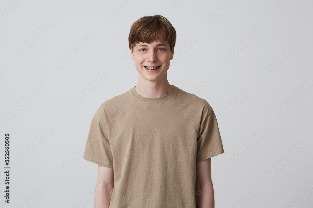 Closeup of cheerful blond handsome young man with healthy teeth and metal braces wears beige t shirt feels happy isolated over white background Looks directly in camera Closeup of cheerful blond handsome young man with healthy teeth and metal braces wears beige t shirt feels happy isolated over white background Looks directly in camera