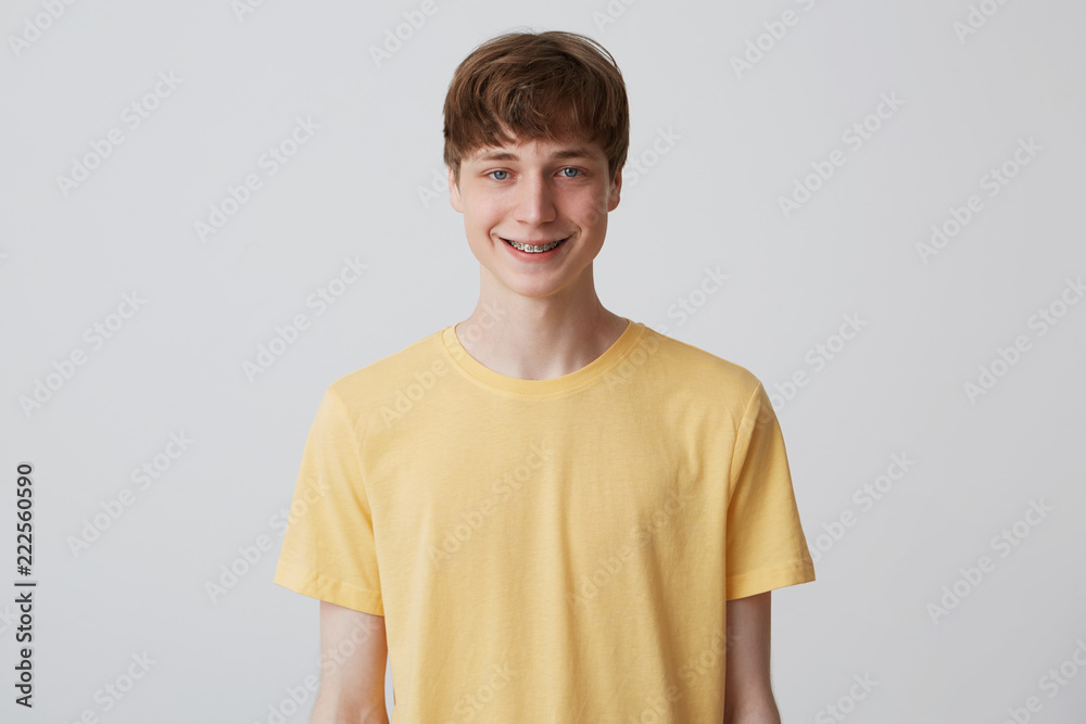Closeup of happy handsome young man with short haircut and braces on teeth wears yellow t shirt standing and smiling isolated over white background Closeup of happy handsome young man with short haircut and braces on teeth wears yellow t shirt standing and smiling isolated over white background