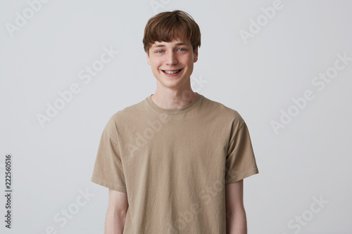 Studio portrait of smiling handsome young man with healthy teeth and metal braces wears beige tshirt and looks directly in camera isolated over white background Feels happy and thinks about girlfriend