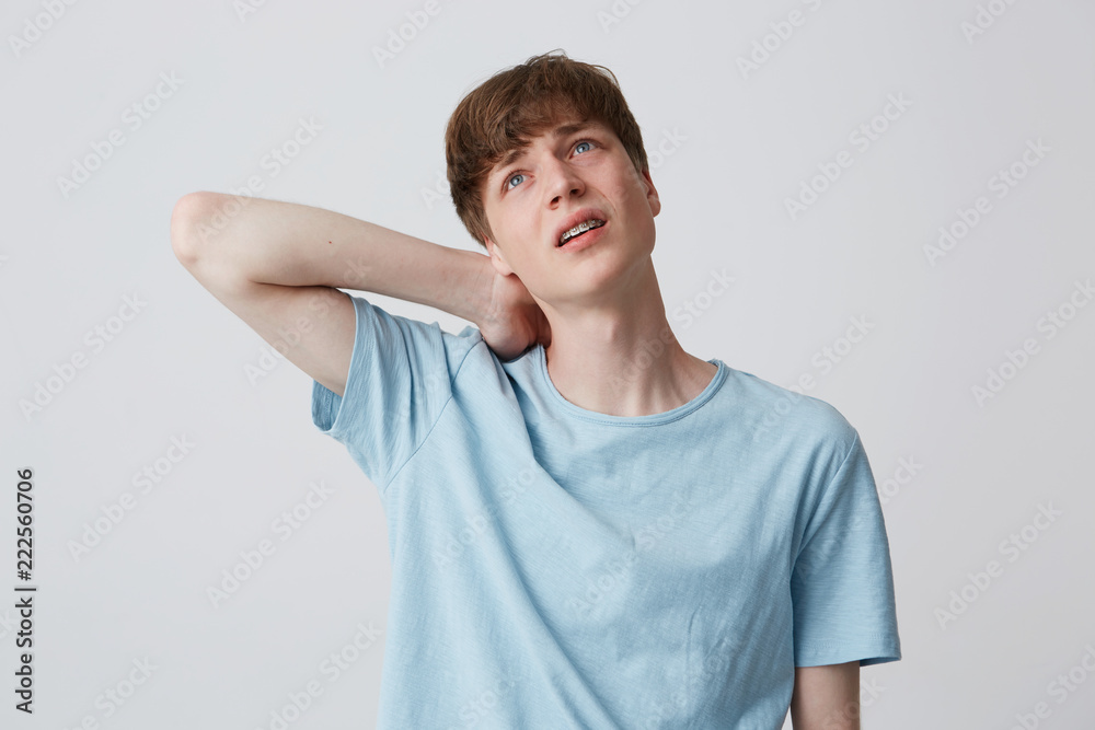 Portrait of sad desperate young man with braces on teeth wears blue t shirt feels tired and suffering from neck pain isolated over white background Portrait of sad desperate young man with braces on teeth wears blue t shirt feels tired and suffering from neck pain isolated over white background