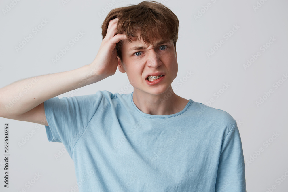 Closeup of angry mad young man with braces on teeth wears blue t shirt keeps hand on head and looks irritated isolated over white background Closeup of angry mad young man with braces on teeth wears blue t shirt keeps hand on head and looks irritated isolated over white background