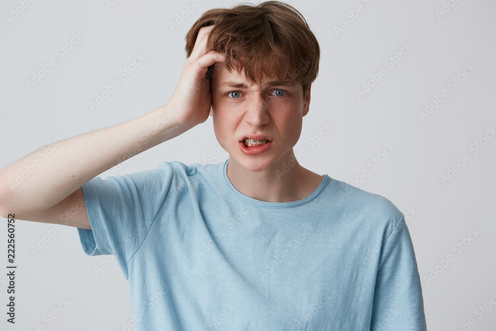 Portrait of stressed displeased young man with braces on teeth wears blue t shirt keeps hand on head and suffering from headache isolated over white background Portrait of stressed displeased young man with braces on teeth wears blue t shirt keeps hand on head and suffering from headache isolated over white background