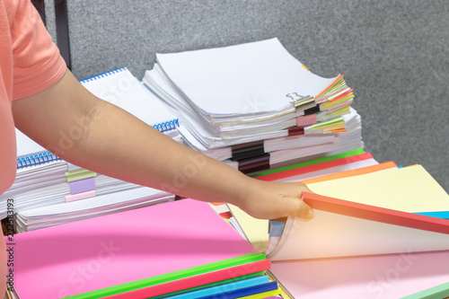 Student is sending homework assignment on teacher's table, which has a large number of paperwork stacked in archive with colorful paper clips and plastic binding bars. Education and business concept.