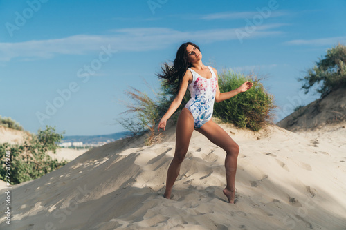 Beautiful young girl posing in a stylish swimsuit on a sandy hill, against the background of wild bushes. It is in an unnatural model position, has a smooth skin, a tan, long dark hair, a beautiful