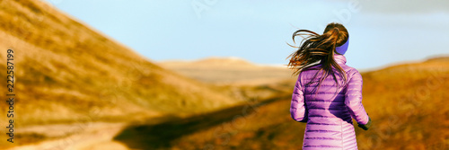 Running woman in autumn foliage background. Athlete runner on trail run outdoors panoramic banner. Active person jogging in down jacket from behind.